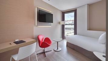 A hotel room with a bed, a desk, a chair, a TV mounted on the wall, and a window with curtains.