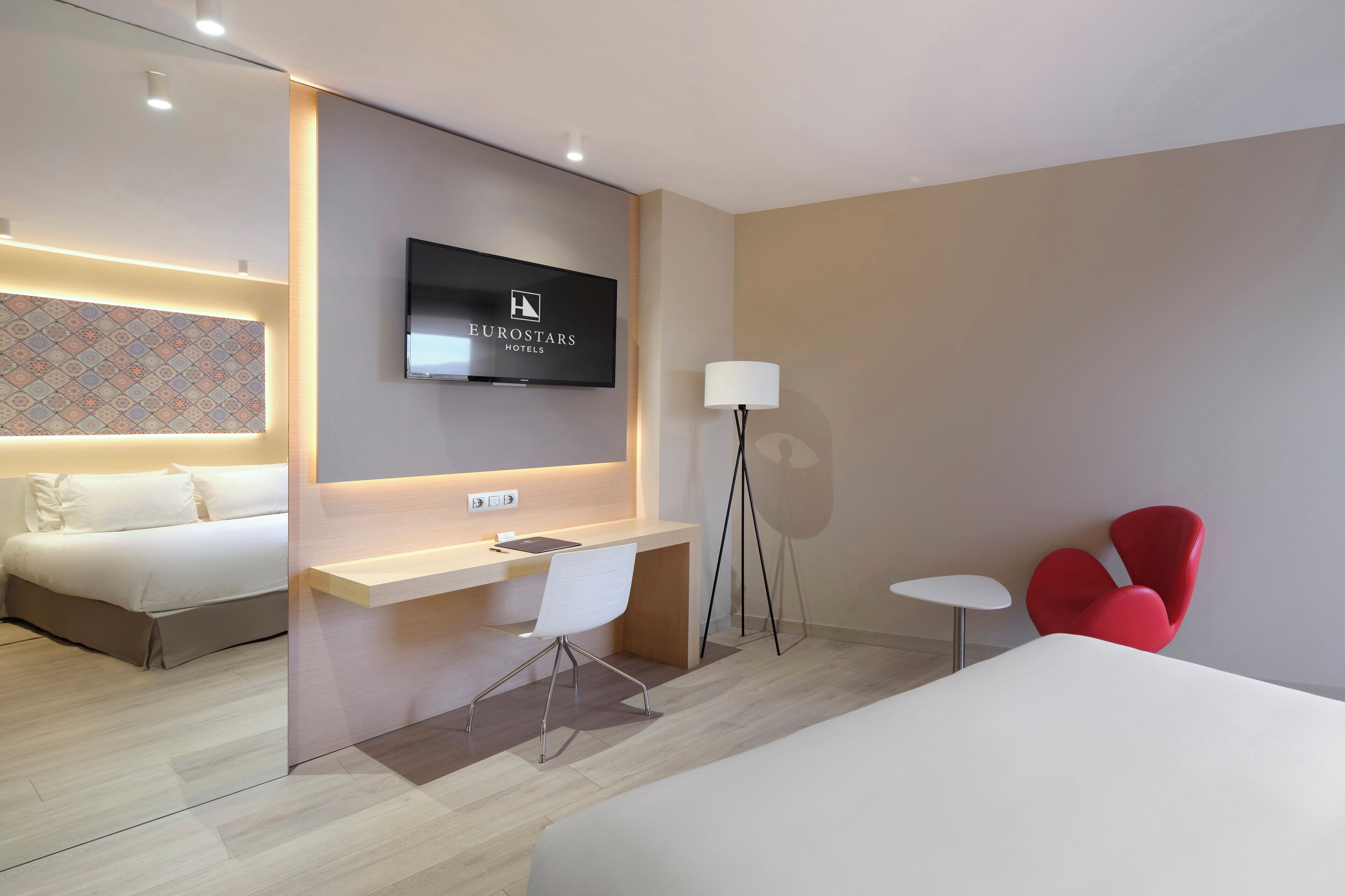 Una camera d'albergo con un letto, una scrivania, una sedia, una televisione, una lampada e una poltrona rossa.