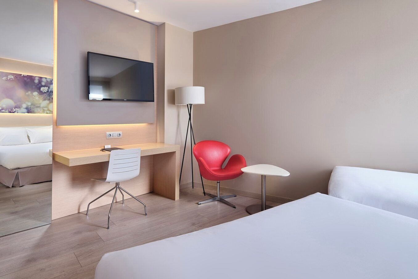 A hotel room with a bed, a desk with a chair, a red chair, a small table, a TV mounted on the wall, and a lamp.