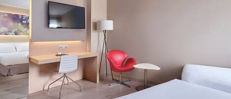 Habitación de hotel con cama, escritorio y silla, una silla roja, una mesita y un televisor montado en la pared, además de una lámpara.