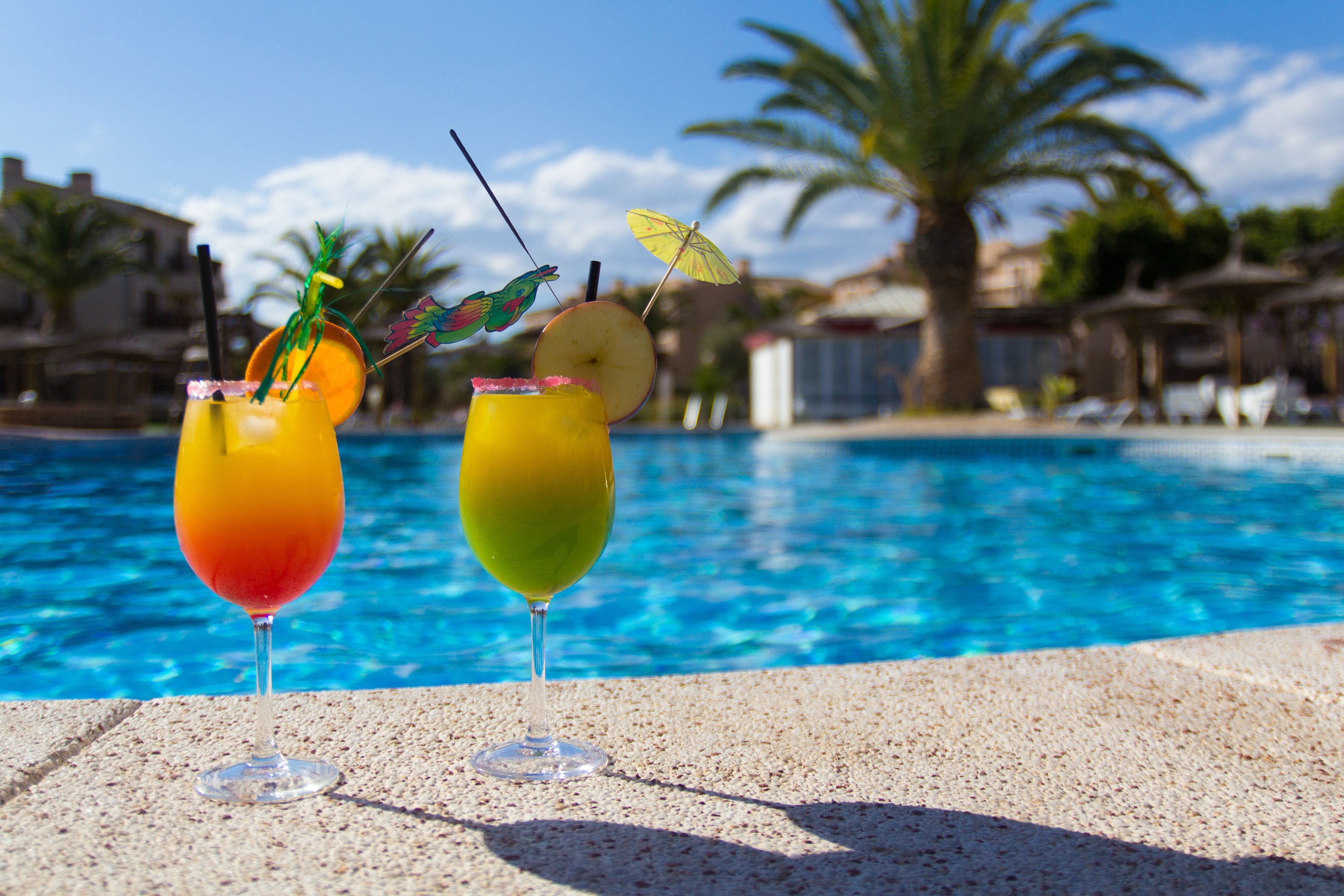 Zwei Cocktails am Pool mit einer Palme und einem Gebäude im Hintergrund.