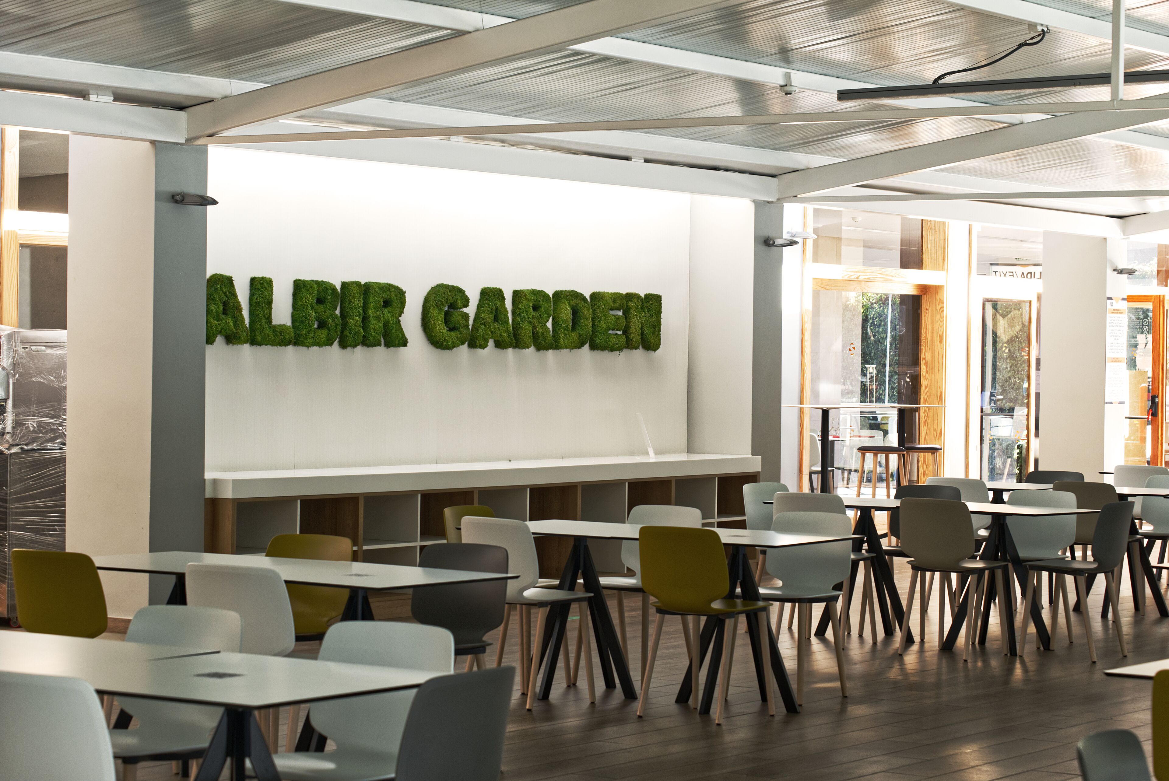 Ein moderner Innenraum mit Tischen und Stühlen, die zum Essen oder für Besprechungen bereitstehen. Eine große Wand zeigt den Text "ALBIR GARDEN".