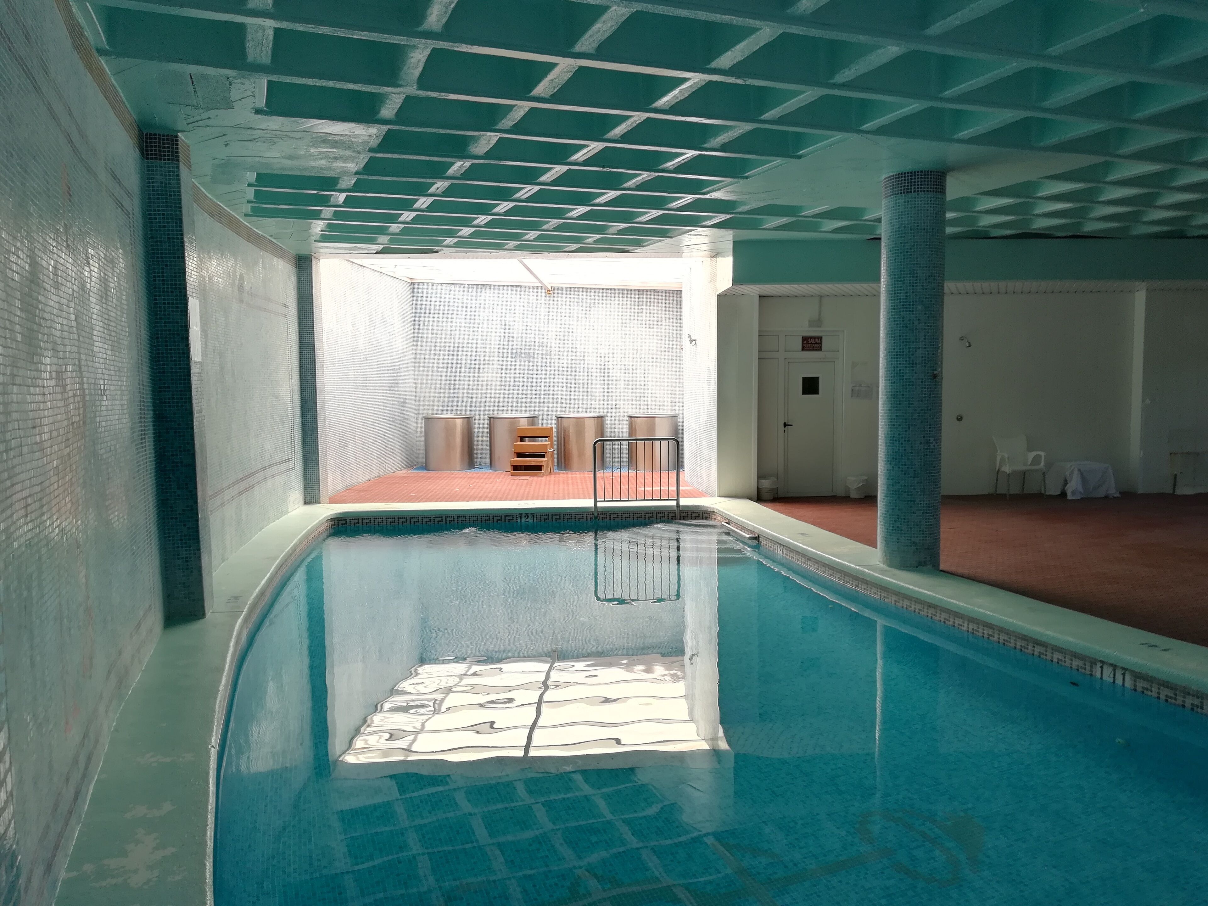 An indoor swimming pool with a tiled floor, a metal ladder, and a white door in the background.