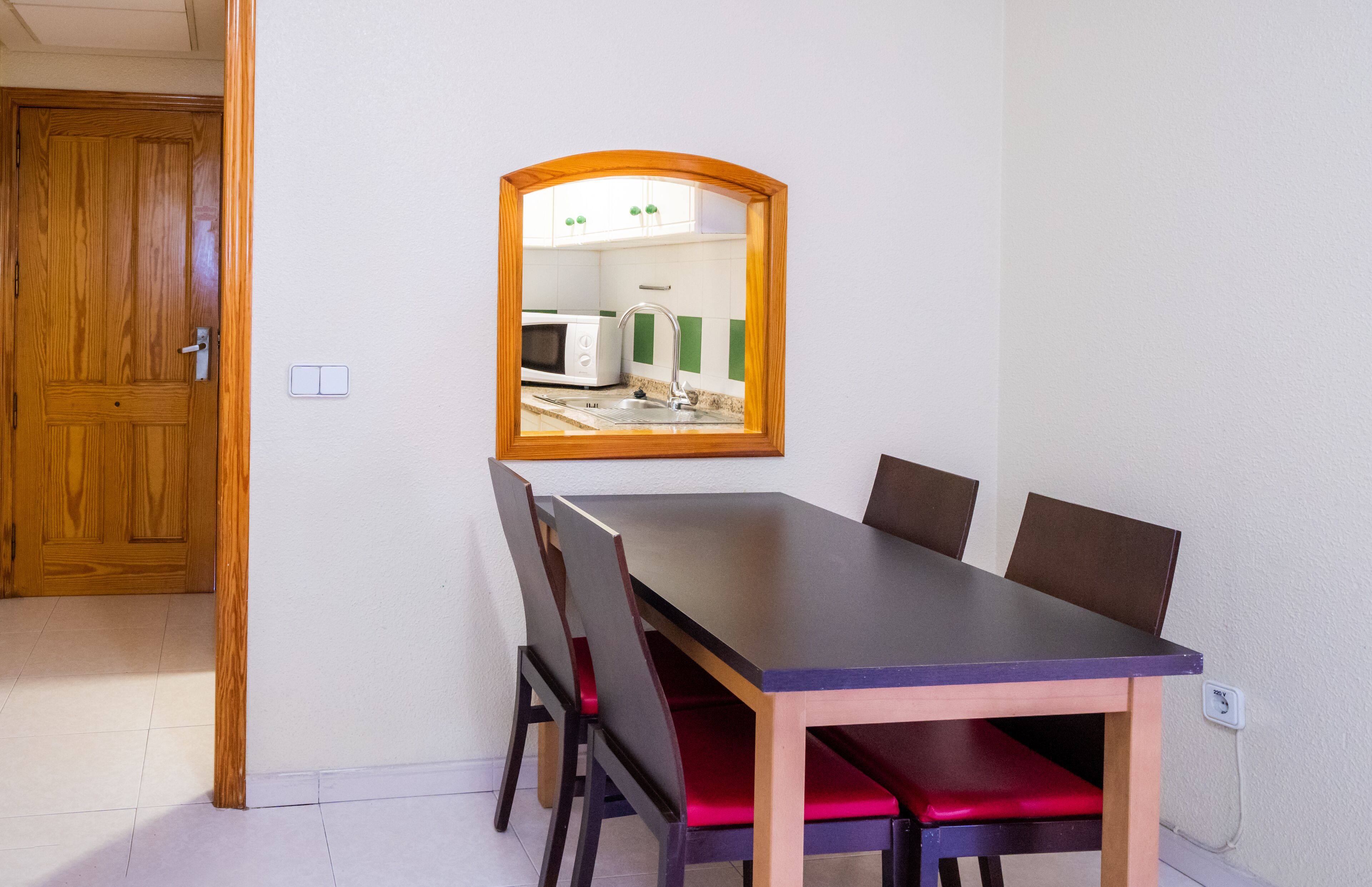 Un comedor con una mesa y sillas de madera, un espejo en la pared y vistas a una cocina con microondas.