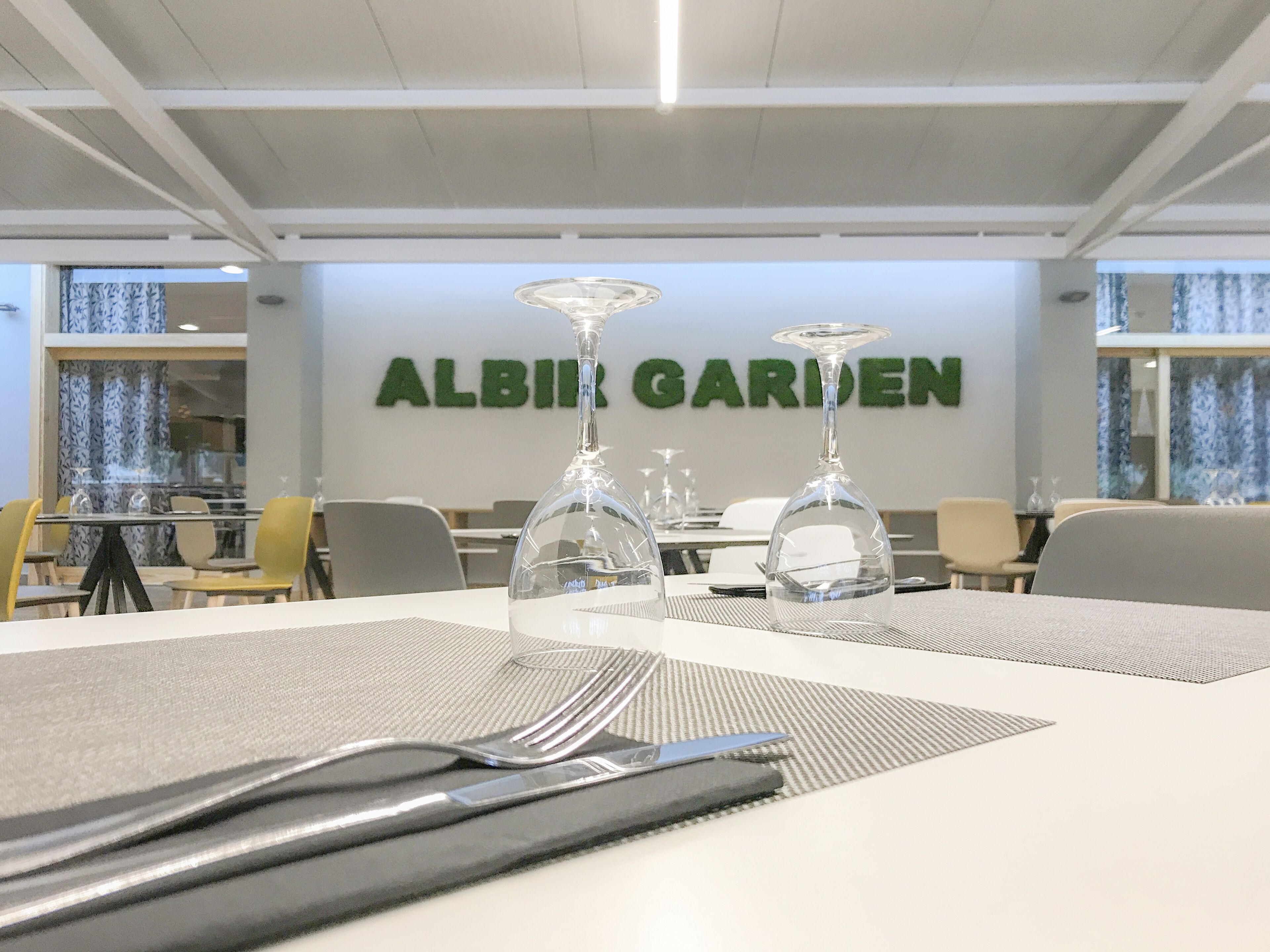 A restaurant with a modern interior, featuring a table set with cutlery and wine glasses, and a sign reading "ALBIR GARDEN" in the background.