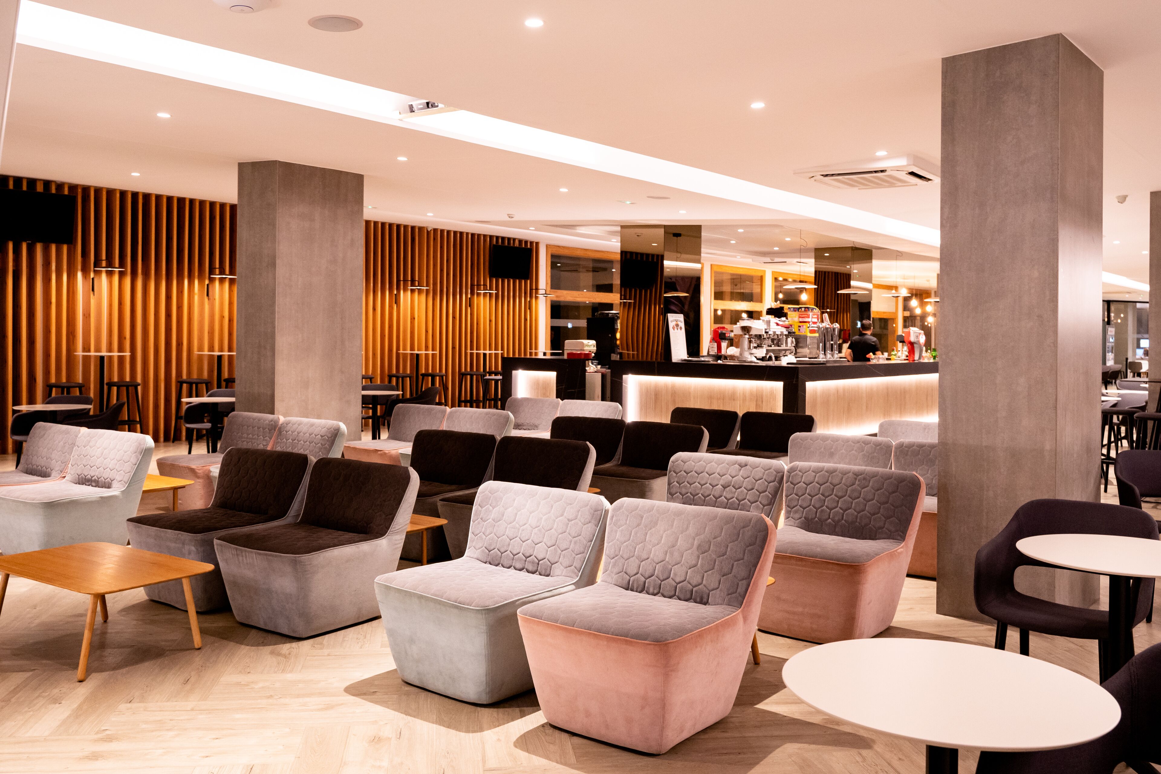 A modern lounge area with sofas and tables, a bar in the background, and wooden paneling on the walls.