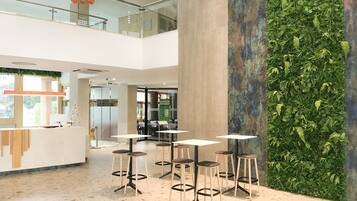 Un intérieur moderne avec un plafond haut, une colonne centrale et un jardin vertical verdoyant. L’espace est aménagé avec plusieurs tables et tabourets.