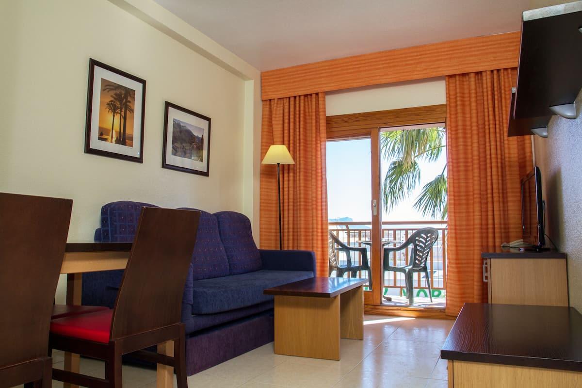 Une pièce comprenant un canapé, des fauteuils, une table, un téléviseur et un balcon avec des rideaux orange.