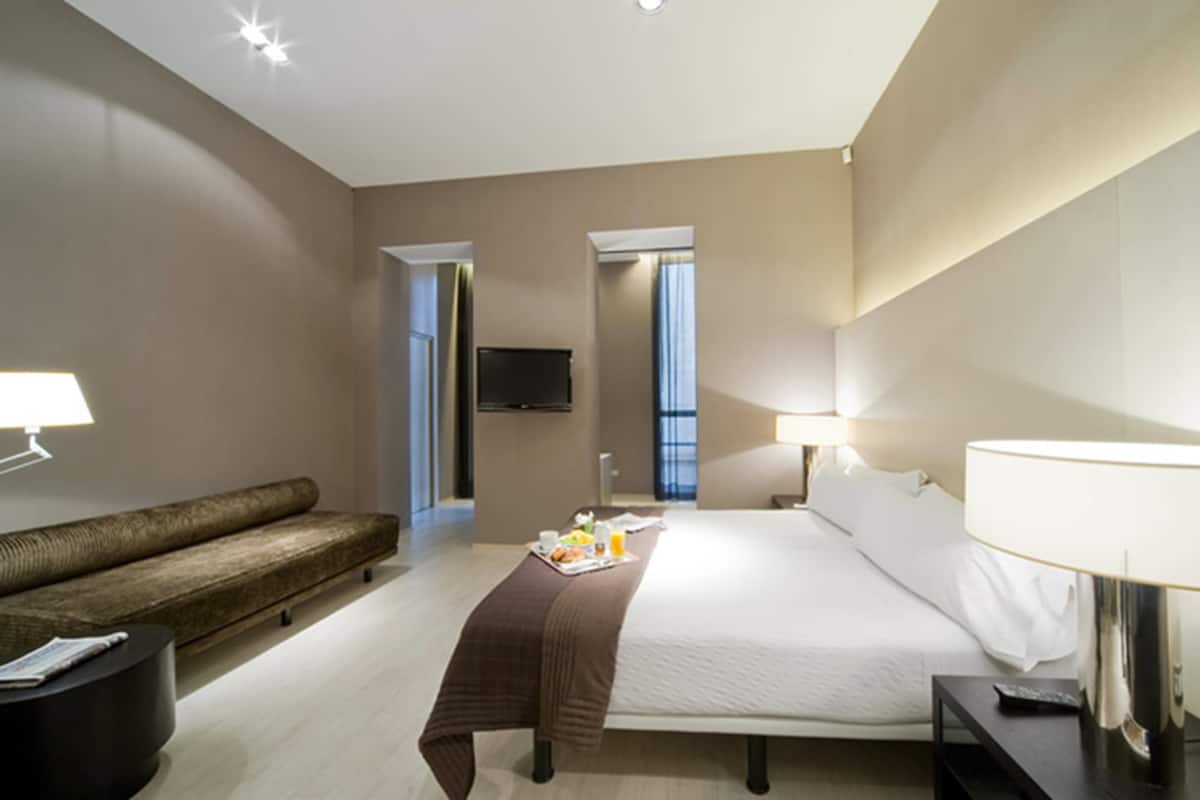 A hotel room with a bed, bedside tables, a sofa, a TV, and a window.