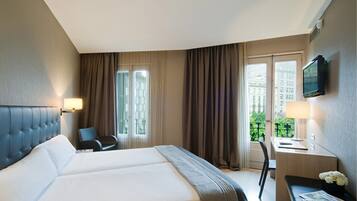 Habitación de hotel con una cama grande, un escritorio con una silla, un televisor montado en la pared y una ventana con cortinas.