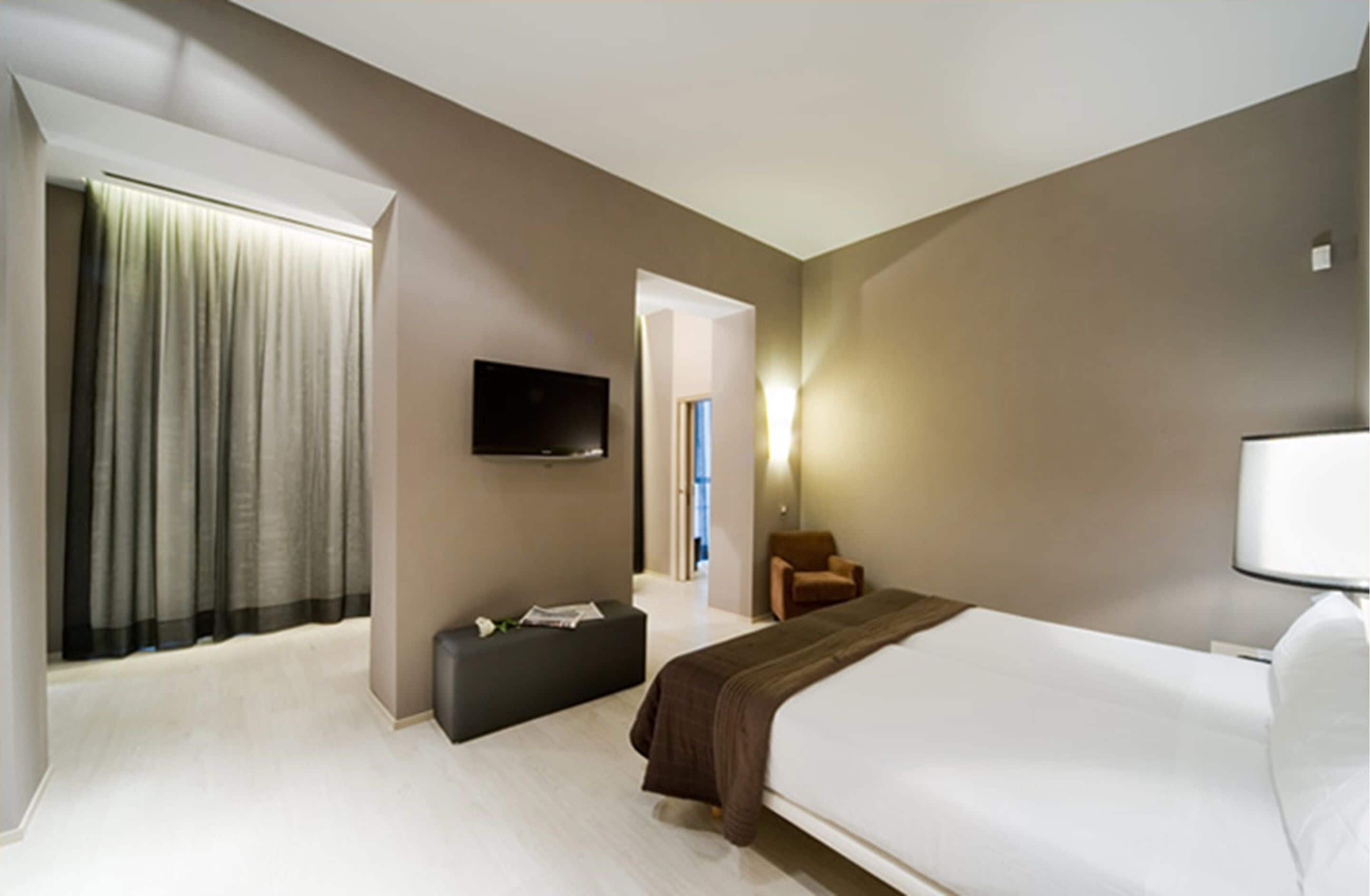 Ein modernes Hotelzimmer mit einem großen Bett, einem flachbildfernseher an der Wand, einer Sitzecke mit einer Bank und einer Tür zu einem weiteren Raum.