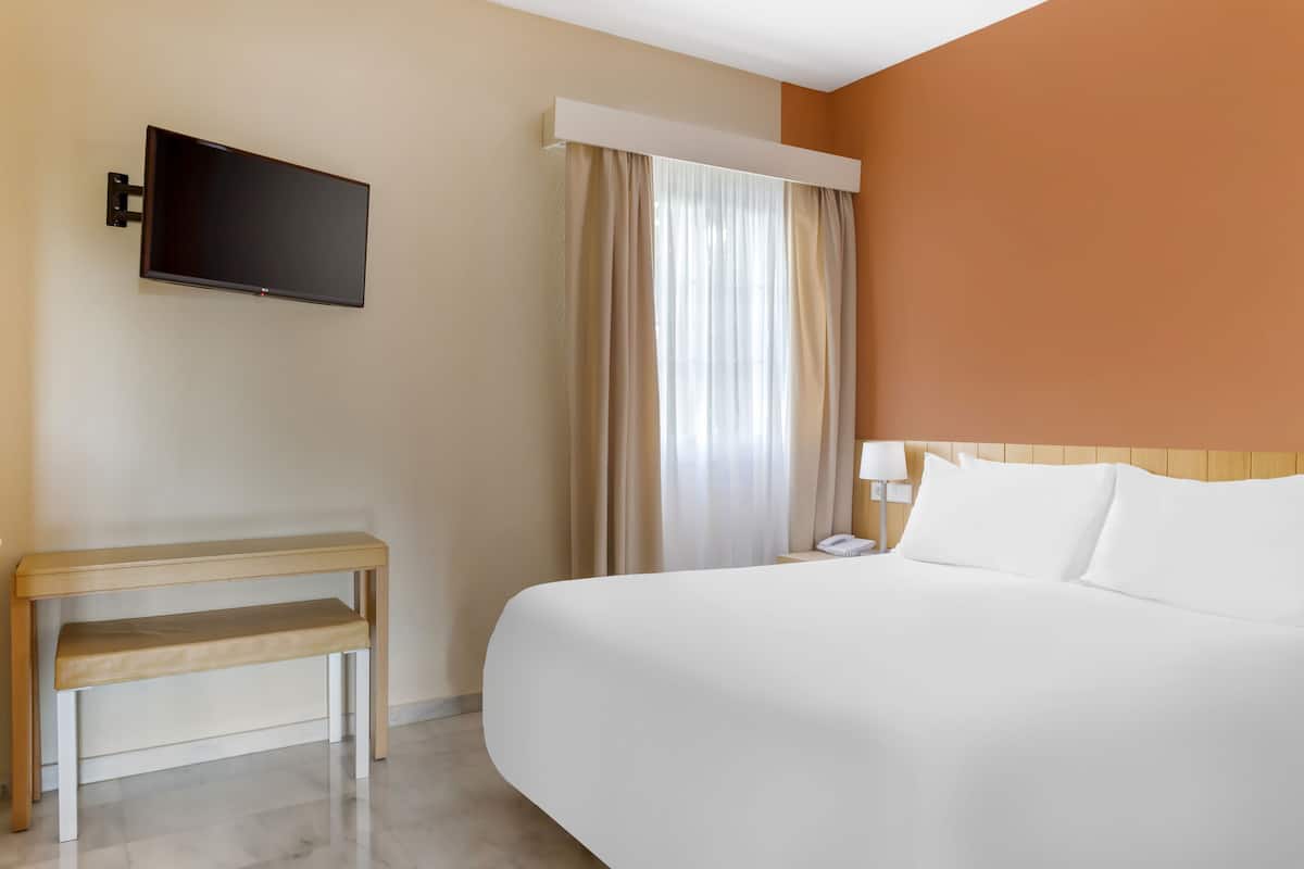 Ein Hotelzimmer mit Bett, Wandfernseher, Nachttisch und einem Fenster mit Vorhängen.