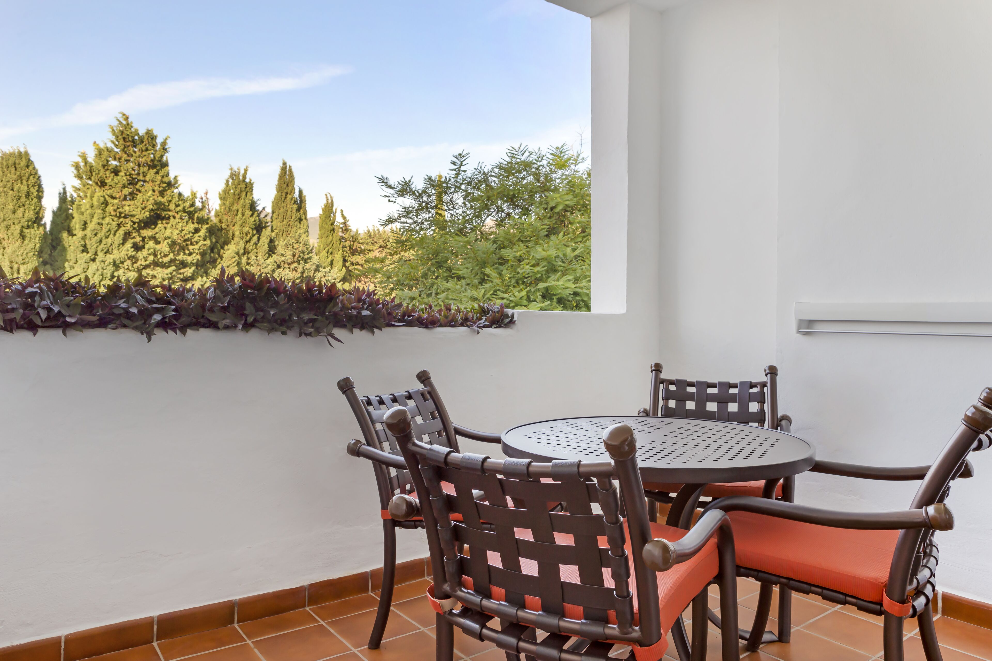 A patio with a round table and four chairs, surrounded by white walls and a view of trees.