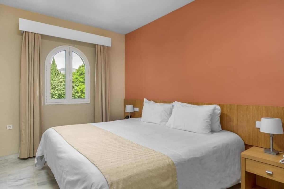 Ein Hotelzimmer mit Bett, Nachttisch mit Lampe, Fenster mit Vorhängen und Blick auf Grünfläche.