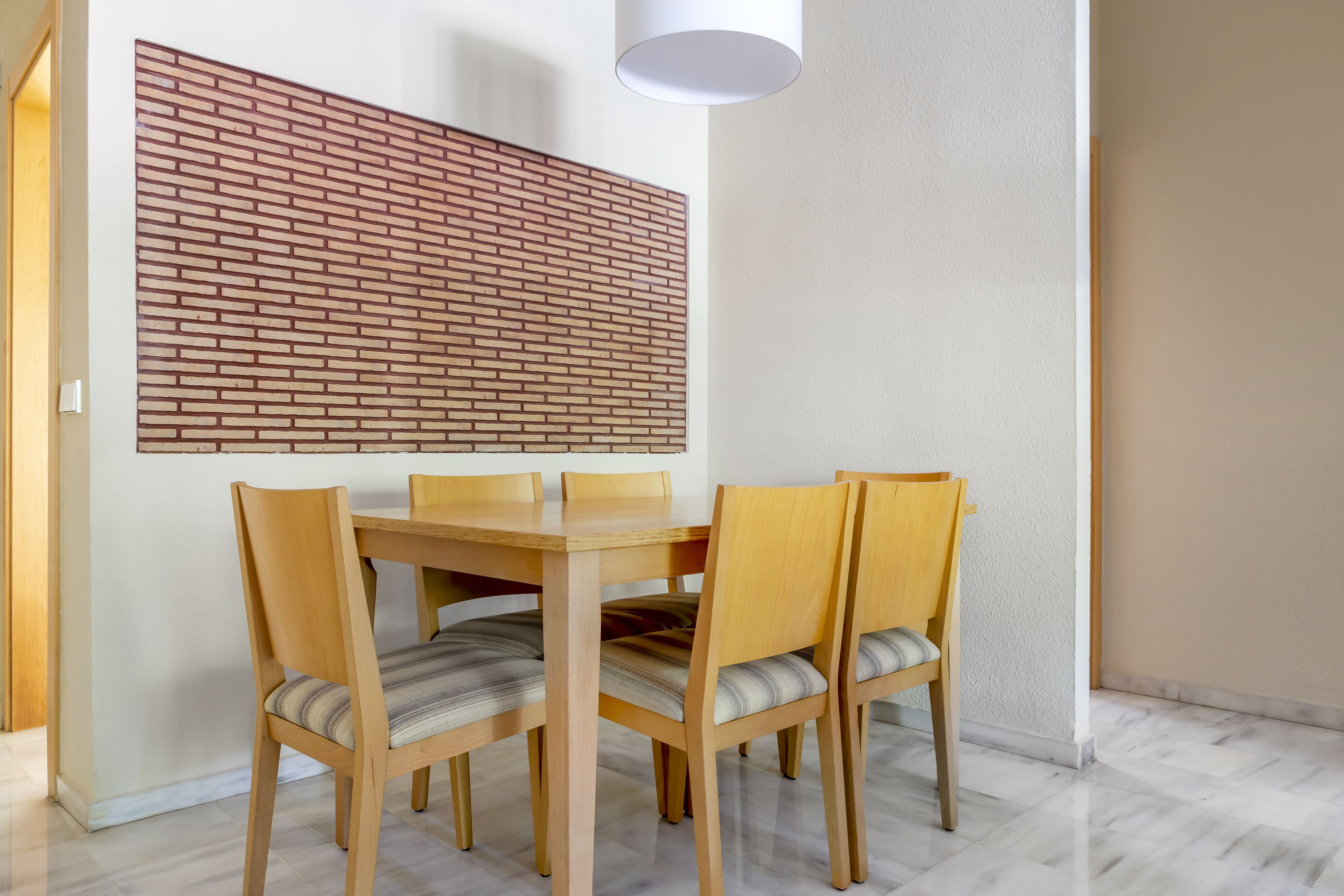 Een eetruimte met een houten tafel en stoelen, een muur met baksteenmotief en een witte plafondlamp.