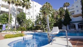 Una piscina con cubierta blanca, rodeada de palmeras y un edificio blanco con balcones.