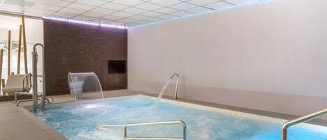 An indoor swimming pool with clear water, a metal ladder, and a wall with a built-in heater.