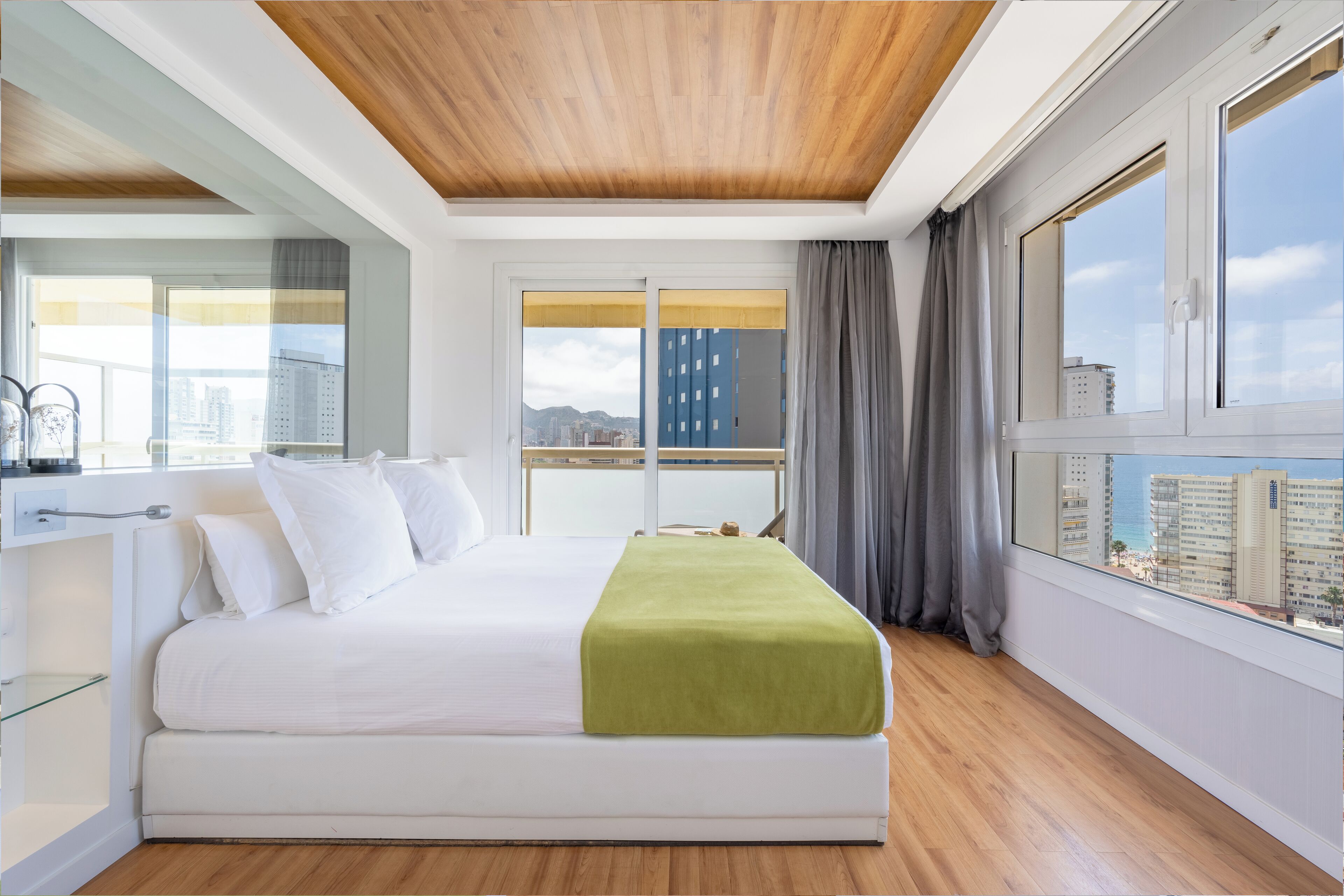 Una habitación de hotel moderna con una cama grande, techo de madera, ventanas amplias con cortinas y vista a la ciudad.