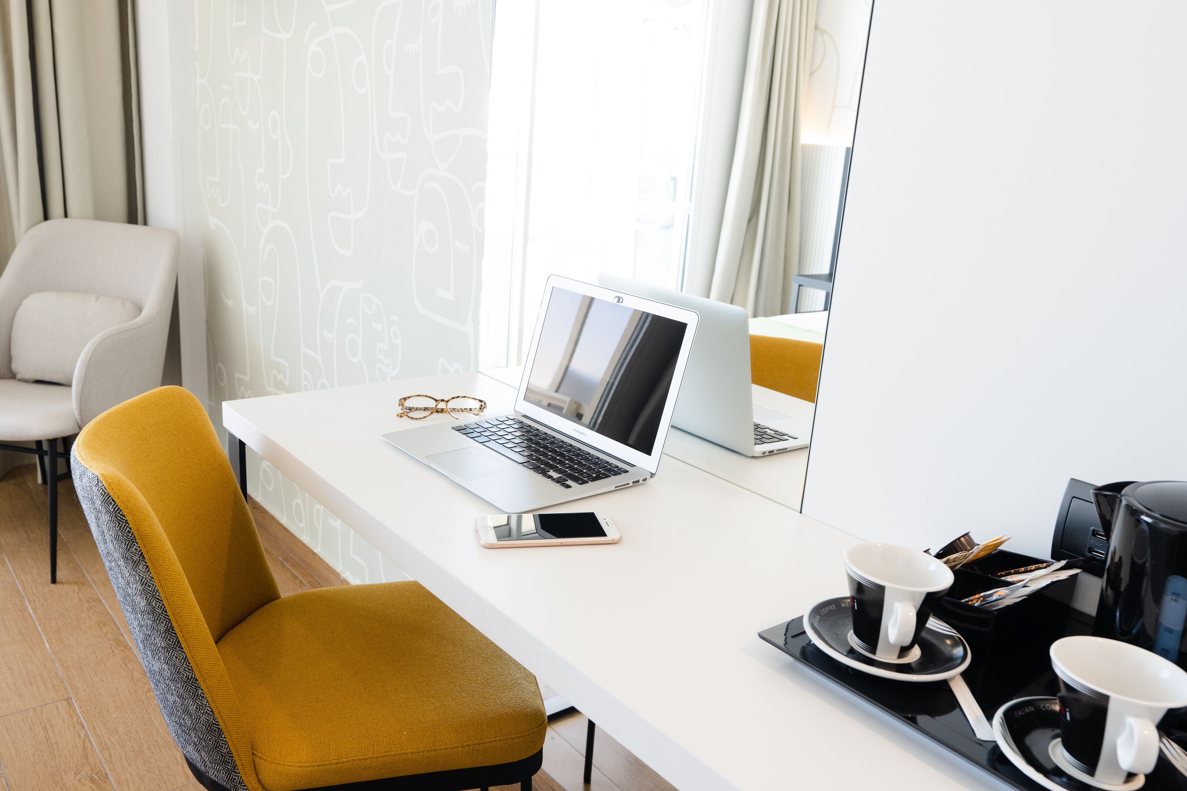 Una habitación de hotel moderna con un escritorio blanco, una computadora portátil, un teléfono inteligente, dos tazas de café y una cafetera.