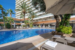 Una piscina con sillones de playa y sombrillas, rodeada de palmeras y un edificio de varios pisos.