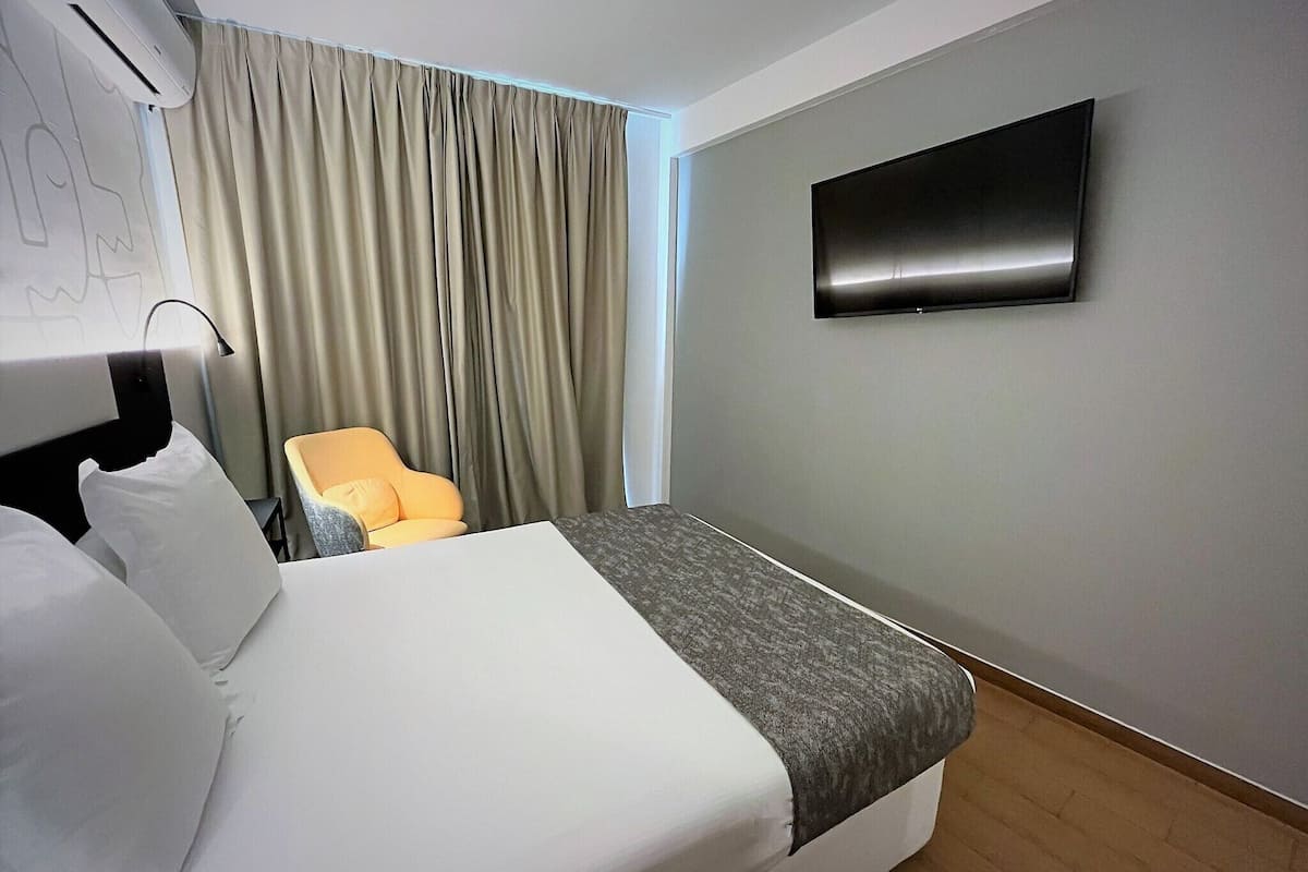 Una habitación de hotel con cama, televisión montada en la pared, una silla y ventana con cortinas.