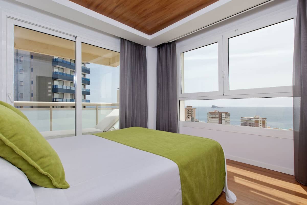 Habitación de hotel con cama, vista al mar y balcón con un edificio al fondo.