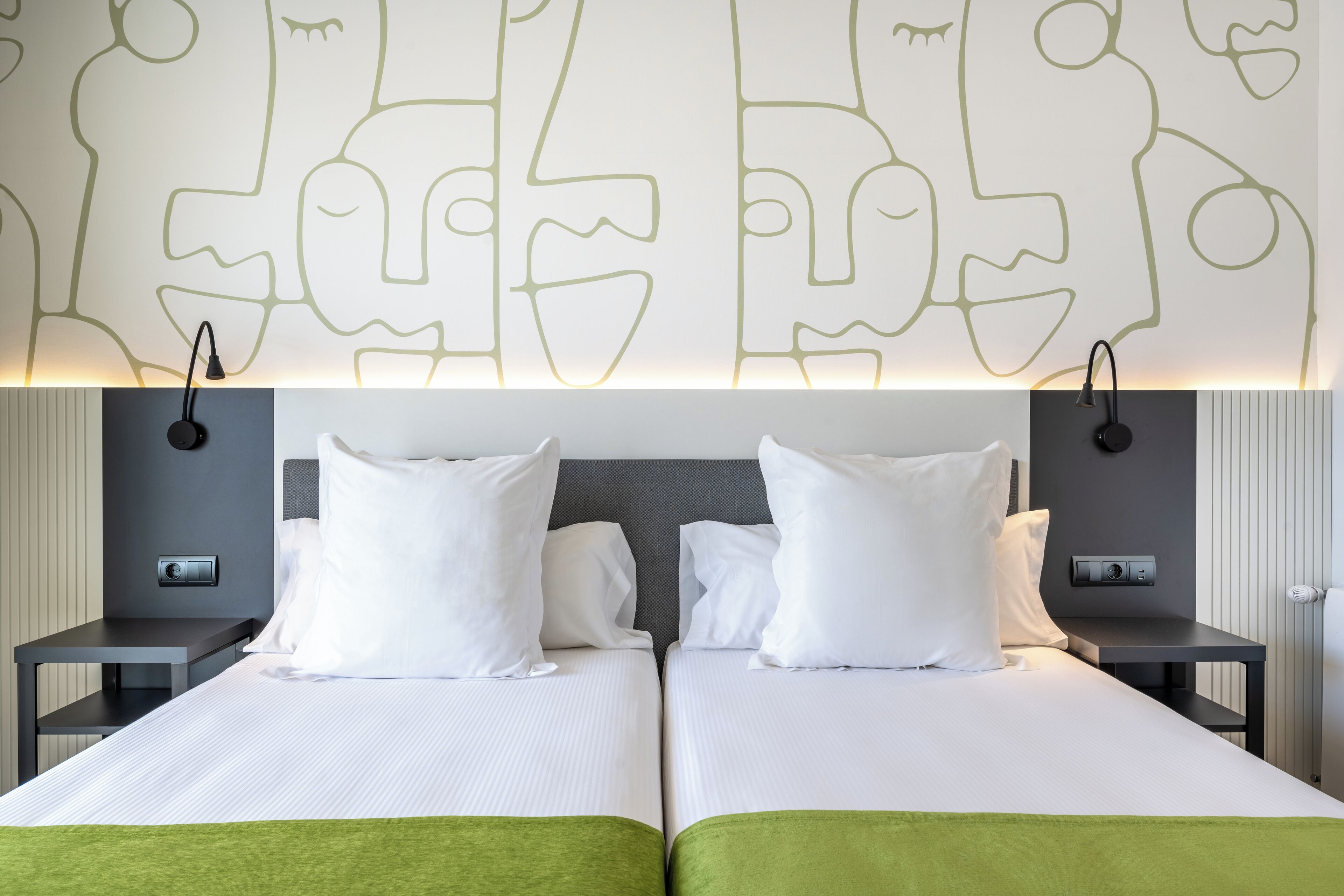 Habitación de hotel con dos camas, una manta verde, un diseño de arte en la pared y una mesita de noche con lámpara.