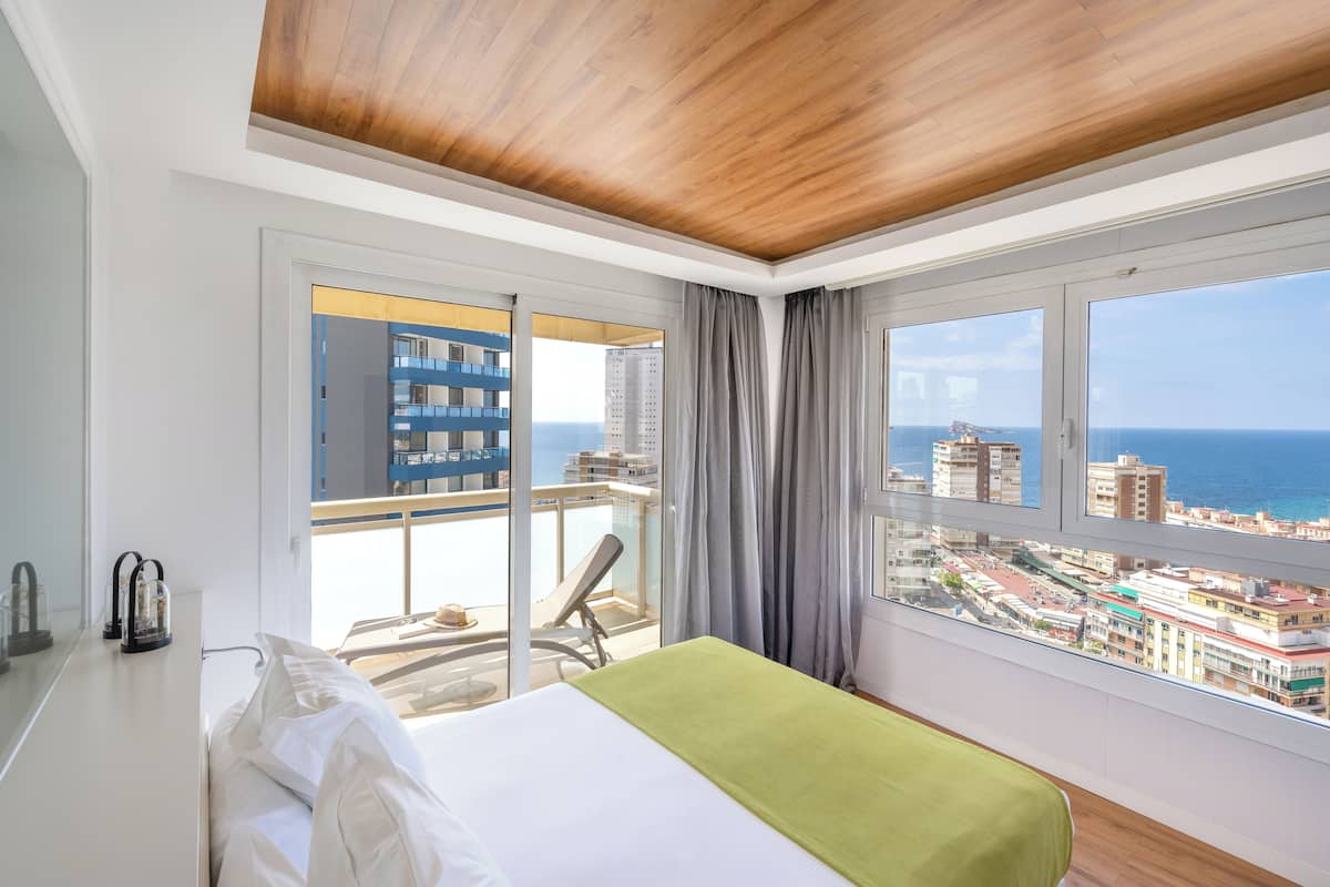 Un cuarto de hotel con cama, una manta verde, vista a la ciudad y al mar, un balcón con una silla y techo de madera.