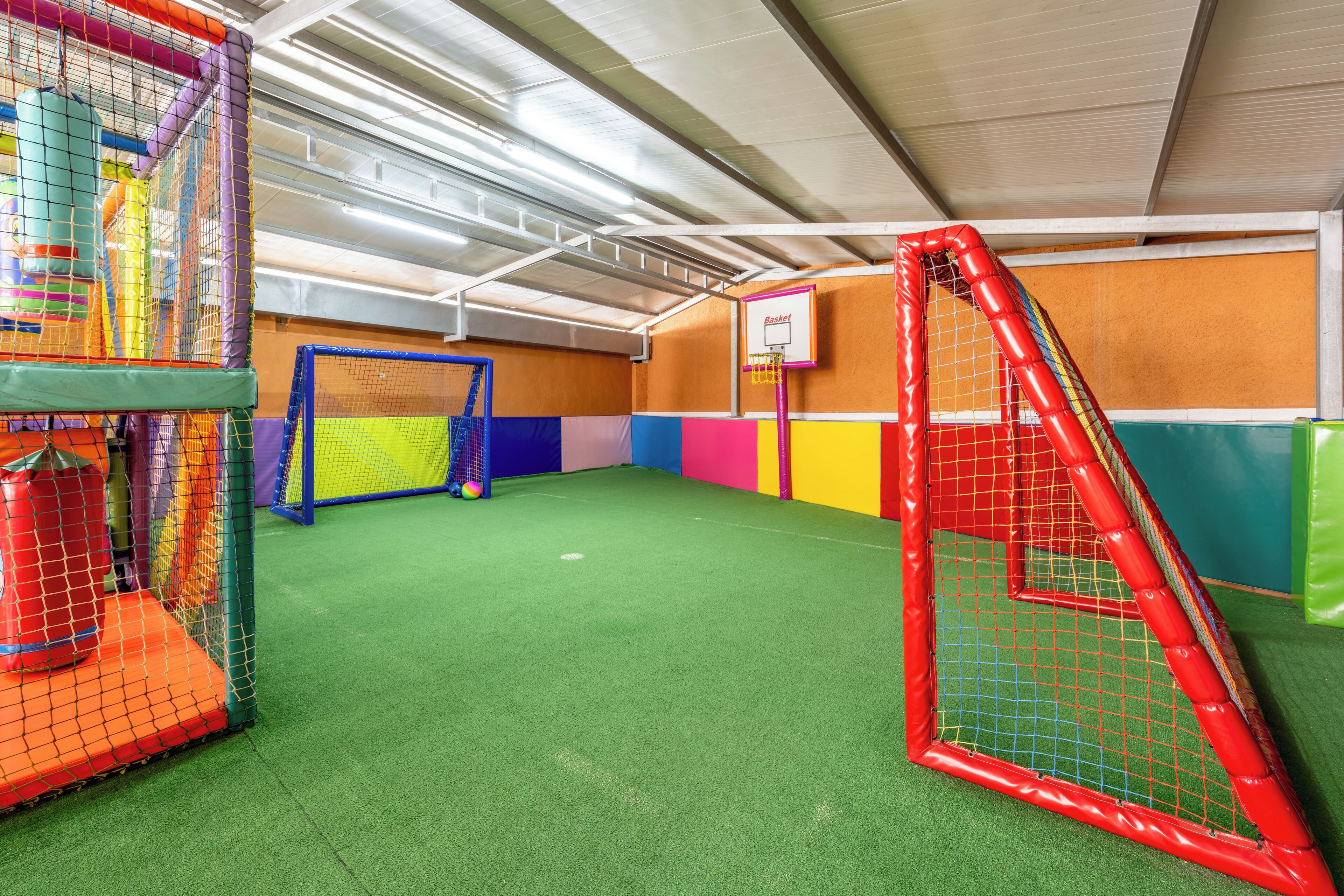Ein Spielbereich im Innenbereich mit Basketballkorb, bunten Wänden und einem grünen Rasenteppichboden.