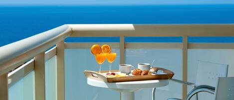A balcony with a tray of breakfast items, including orange juice, croissants, and a cup of coffee, overlooking a clear blue sea.