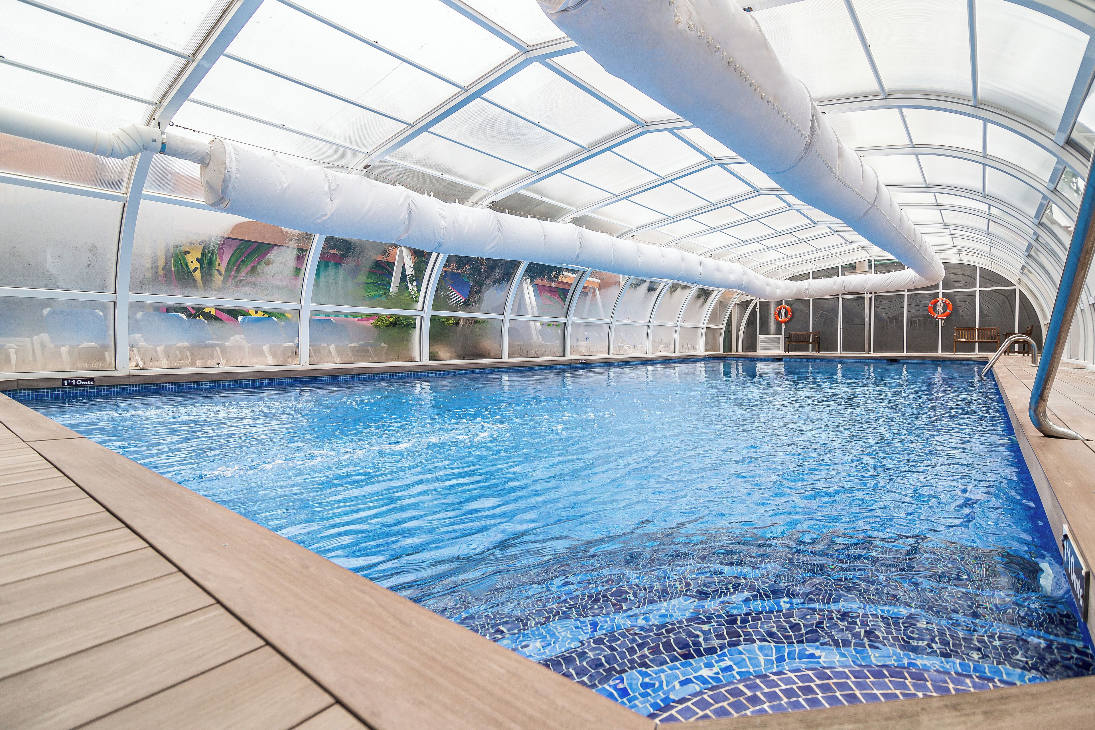 An indoor swimming pool with clear blue water, surrounded by a wooden deck and a glass ceiling structure.
