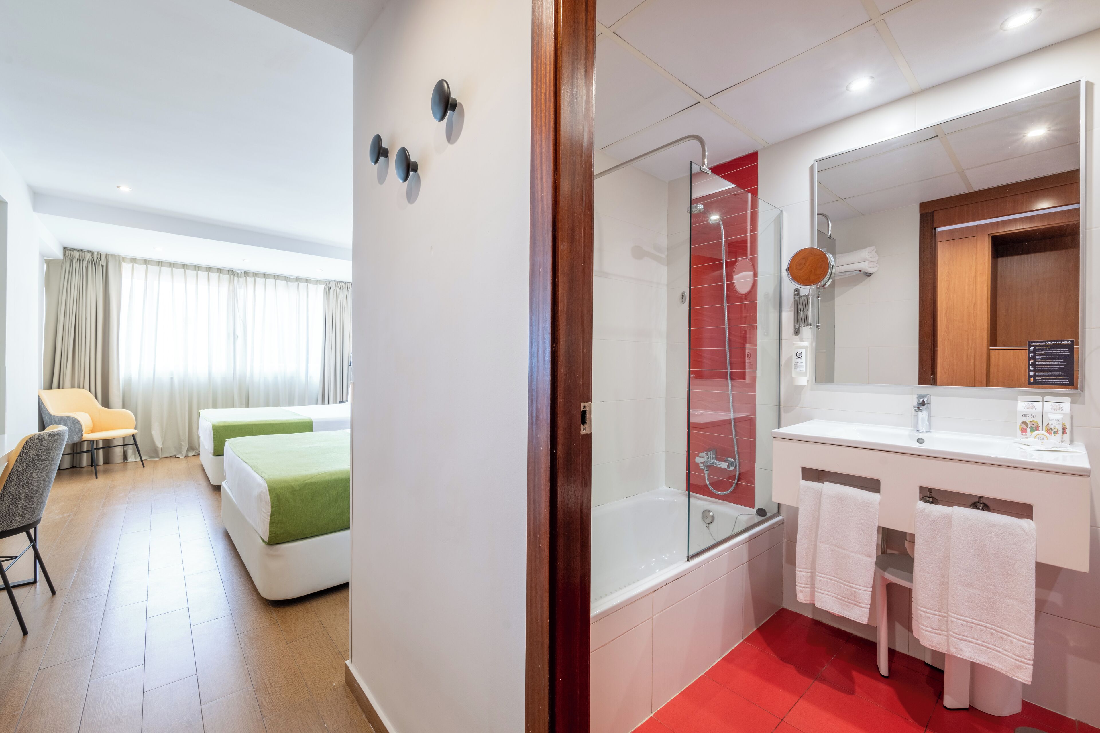 Quarto de hotel com cama, escrivaninha, cadeira, chuveiro e banheiro com pia e espelho.