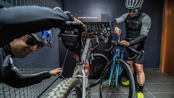 Dos personas ajustando bicicletas en una habitación con piso de baldosas y un perchero para bicicletas en la pared.