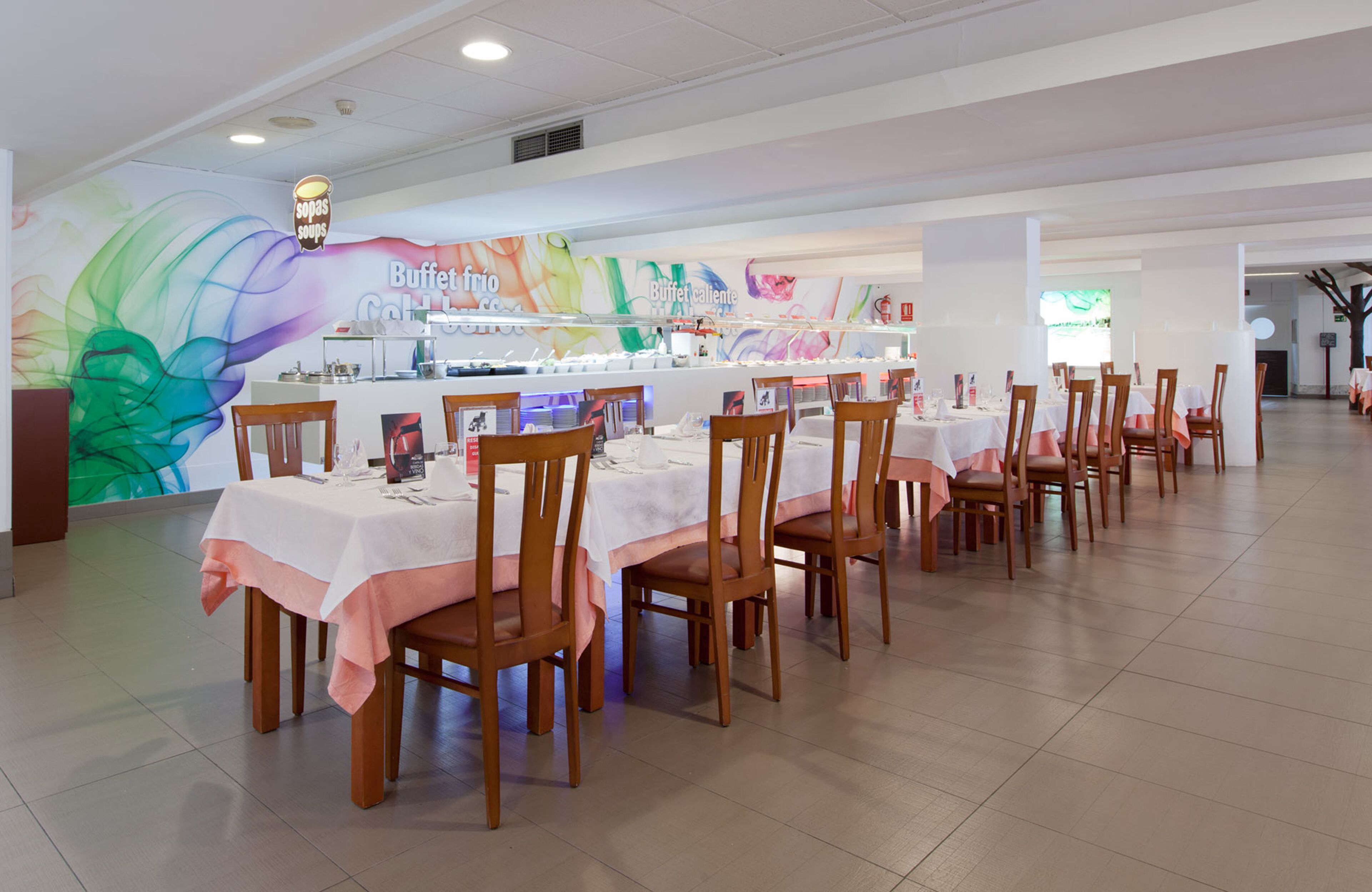A restaurant with multiple tables and chairs, a colorful mural on the wall, and a buffet area.