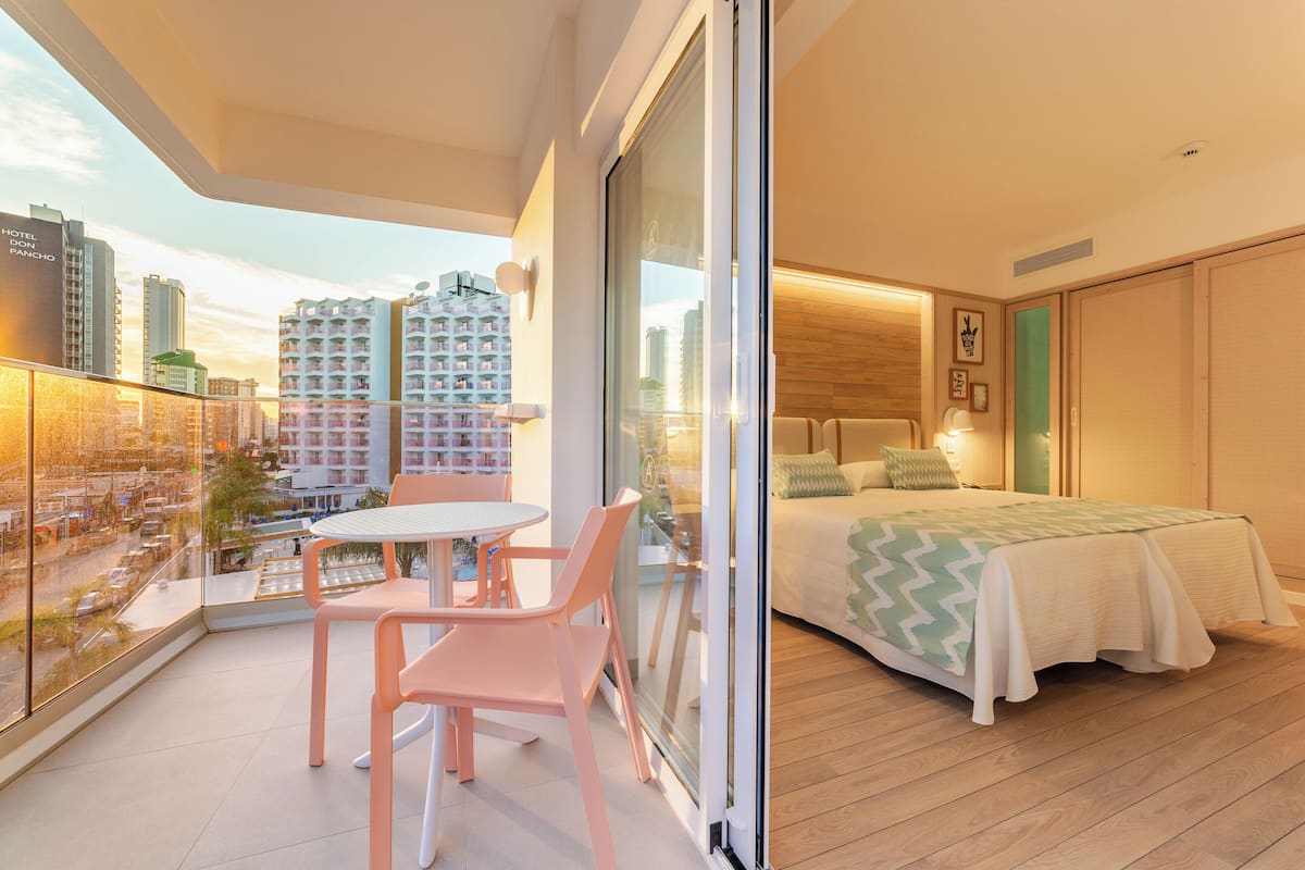 A modern hotel room with a balcony, a bed with a green zigzag bedspread, and a view of high-rise buildings.