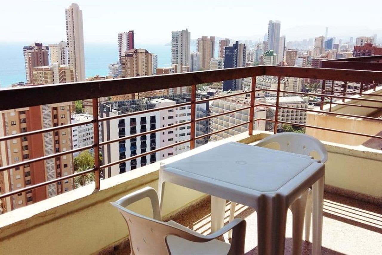 Un balcón con mesa y sillas, con vista a un paisaje urbano de edificios altos y al mar.