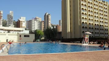 Zona de piscina con un puesto de socorrista, sillas junto a la piscina y una vista de un alto edificio de hotel al fondo.