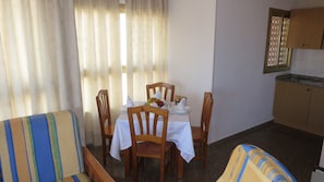 Uma área de refeições com uma mesa posta para quatro pessoas, cadeiras de madeira, um sofá listrado e uma cozinha com armários.