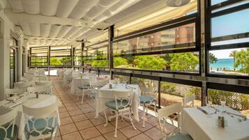 Un restaurant doté d’une terrasse, de tables et de chaises blanches, et de grandes fenêtres offrant une vue sur la mer et les bâtiments.