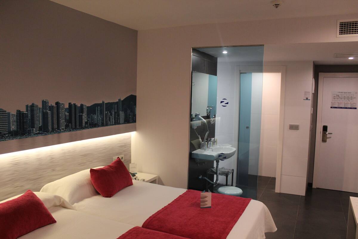 Quarto de hotel com cama, almofadas vermelhas, um mural com vista da cidade, banheiro com pia e espelho, e porta.