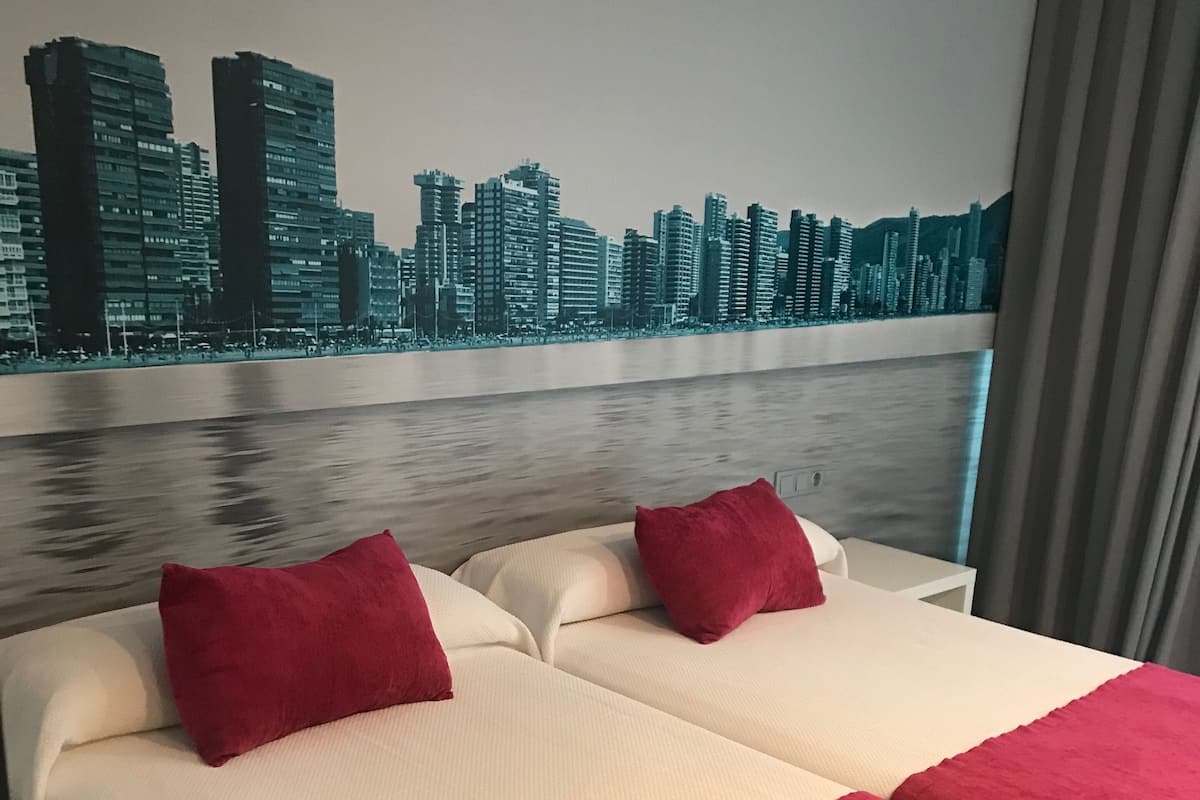 Quarto de hotel com duas camas, cada uma com almofadas vermelhas e colcha branca. Um mural com vista urbana está na parede atrás das camas, e uma janela com cortinas é visível.