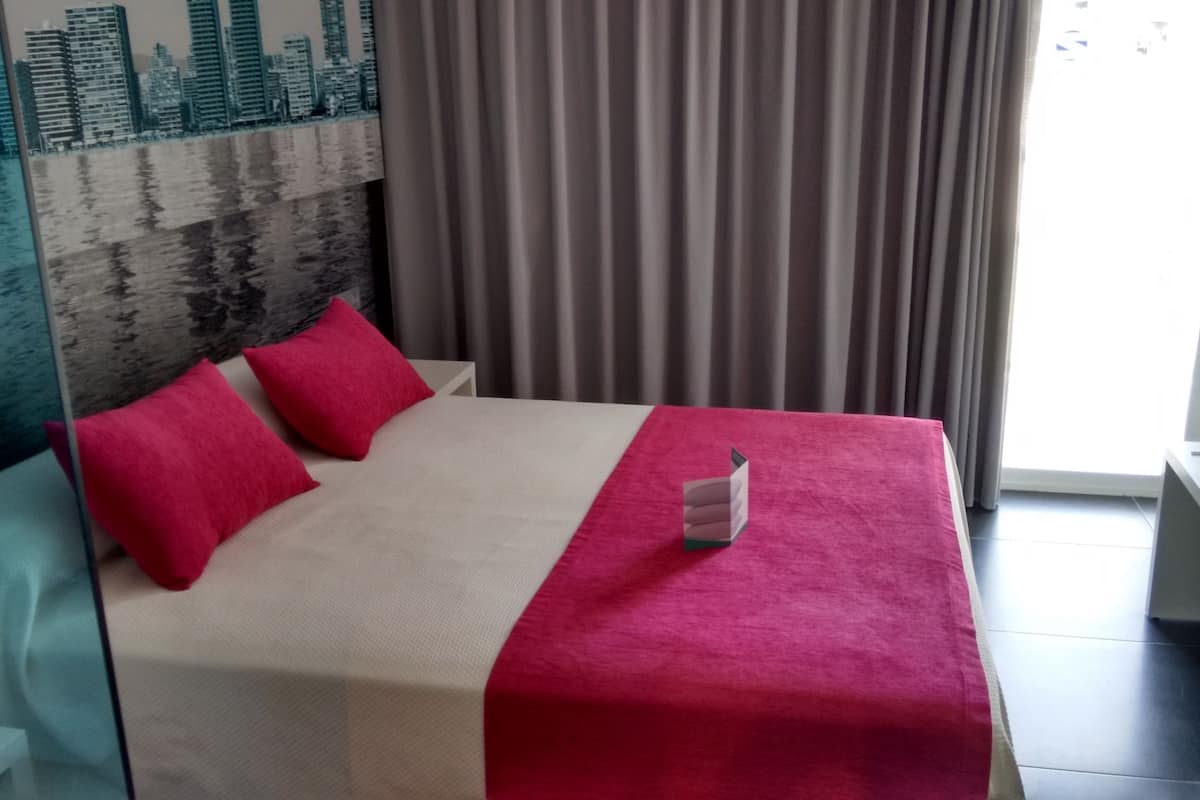 Quarto de hotel com cama, almofadas e manta vermelhas, painel de parede com vista urbana e janela com cortinas.