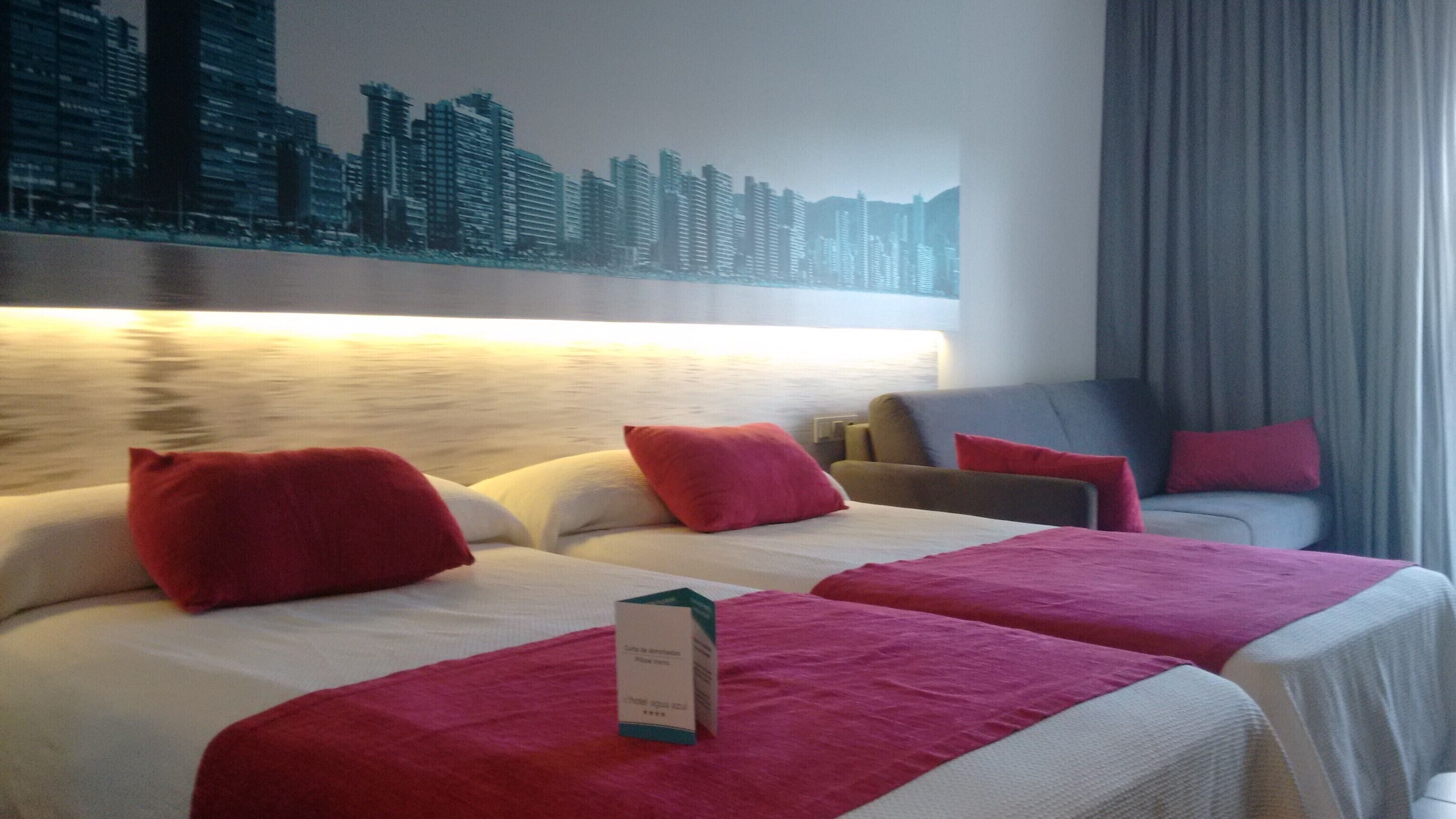 Habitación de hotel con dos camas, almohadas rojas, un mural de paisaje urbano, un sofá y una cortina.