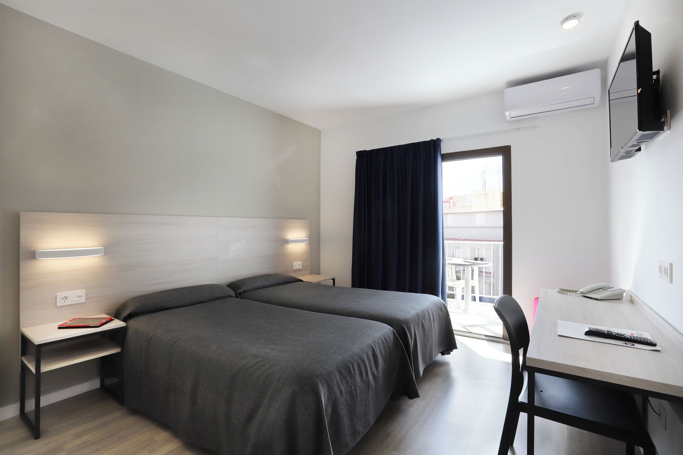 Quarto de hotel com duas camas, uma escrivaninha com cadeira, TV fixada na parede e janela com cortinas.