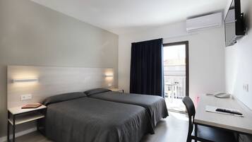 Quarto de hotel com duas camas, uma escrivaninha com cadeira, TV fixada na parede e janela com cortinas.