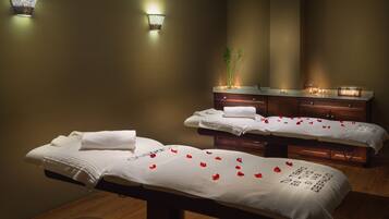 A spa room with two massage tables, each with a towel and rose petals. There are wall-mounted lights and a wooden cabinet with a mirror above the tables.