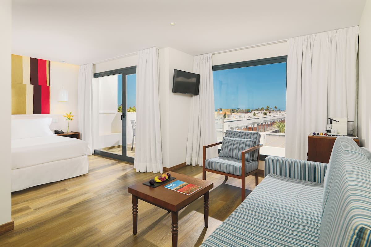 A hotel room with a sofa, a chair, a table with a book and fruit, a TV, and a balcony with a view of the city.