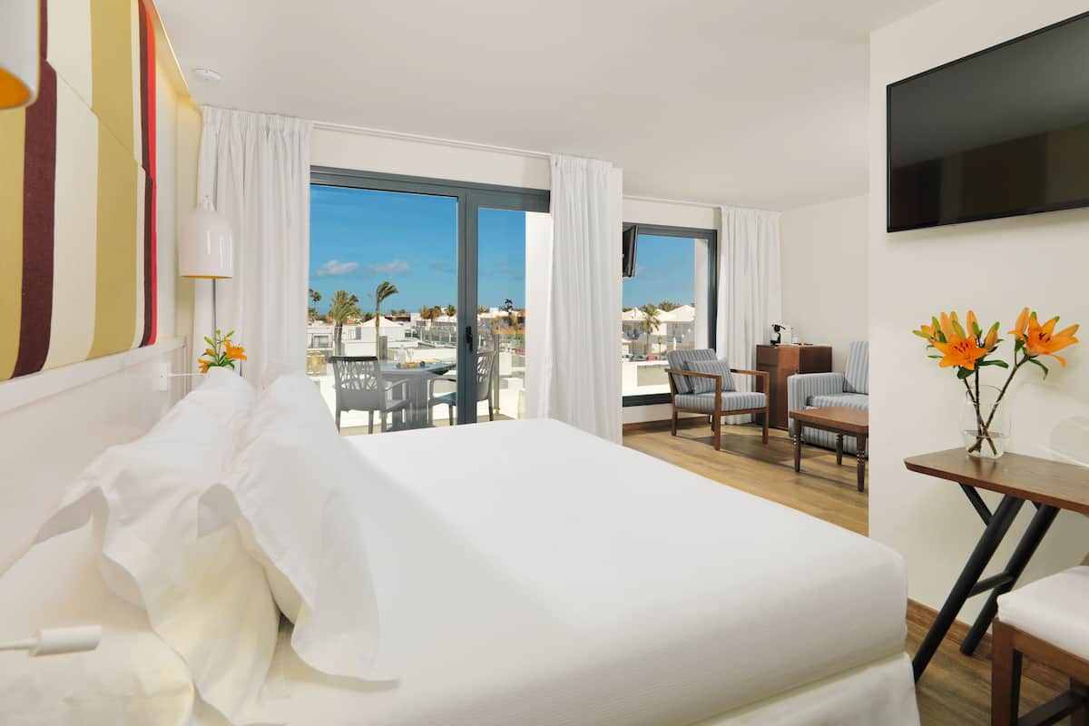 A hotel room with a large bed, a balcony with a view of the resort, a TV, and a table with flowers.