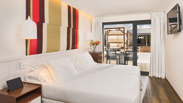 A hotel room with a large bed, a bedside table with a lamp, a desk with a laptop, and a view of the outdoor seating area through a glass door. - H10 Ocean Dreams Boutique Hotel - Adults Only (La Oliva)