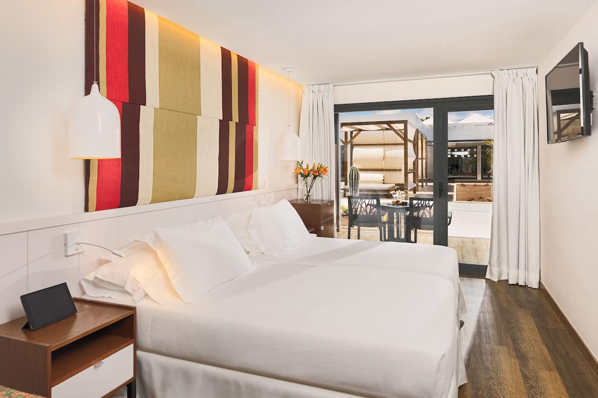 A hotel room with a large bed, a bedside table with a lamp, a desk with a laptop, and a view of the outdoor seating area through a glass door.